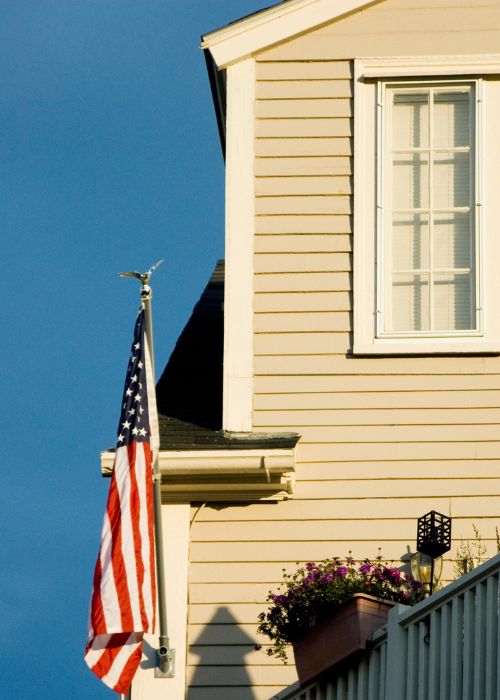 Image of a beige Connecticut home with an American flag and blue sky on  the About CT Deck and Gutter page.